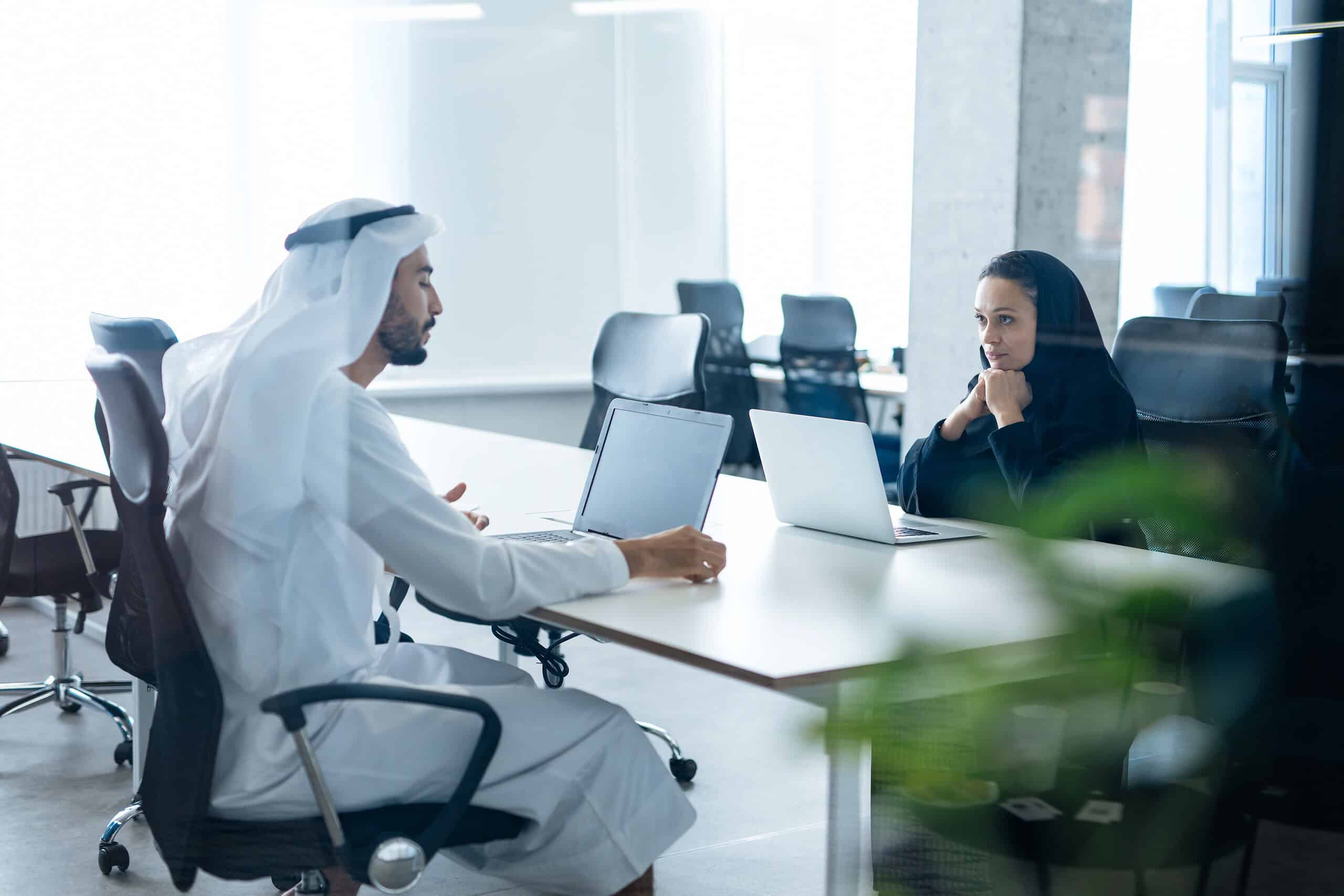 Man and woman with traditional clothes working in a business office of Dubai. Portraits of successful entrepreneurs businessman and businesswoman in formal emirates outfits. Concept about middle east