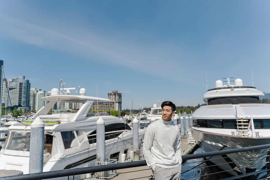 Portrait of asian rich man standing near expensive yachts, looking away outdoors. Attractive chinese male posing for picture, on street