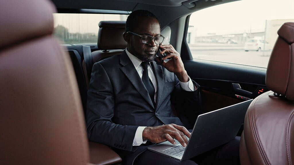 Portrait of successful serious African American businessman traveling by car talking on phone and typing on laptop. Influential attractive African man in suit having business trip.