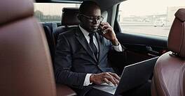 Portrait of successful serious African American businessman traveling by car talking on phone and typing on laptop. Influential attractive African man in suit having business trip.
