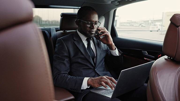 Portrait of successful serious African American businessman traveling by car talking on phone and typing on laptop. Influential attractive African man in suit having business trip.