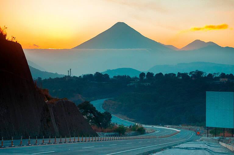 Volcano called "Agua" at sunset in Guatemala Central America, silhouette of mountains and modern highway, dramatic landscape on the road, play of light and shadow.