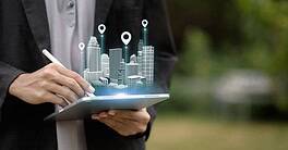 A person uses a tablet to project a holographic 3D model of a smart city with location markers. Concept of urban planning, real estate technology, architecture, and navigation.