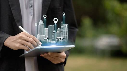 A person uses a tablet to project a holographic 3D model of a smart city with location markers. Concept of urban planning, real estate technology, architecture, and navigation.