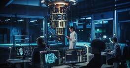 Female Engineer Working on Quantum Computer Hardware, Using Tablet Computer to Access Technical Specifications and Control Processor Parameters. Group of Developers Work on AI Behind a Desk