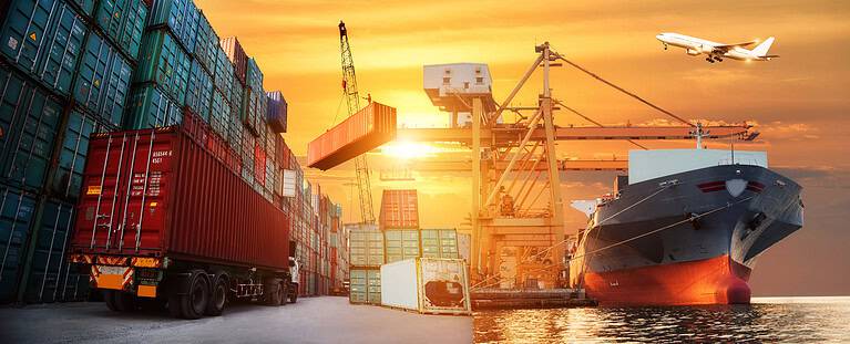Cargo ship, containers, cranes, and airplane at sunset.